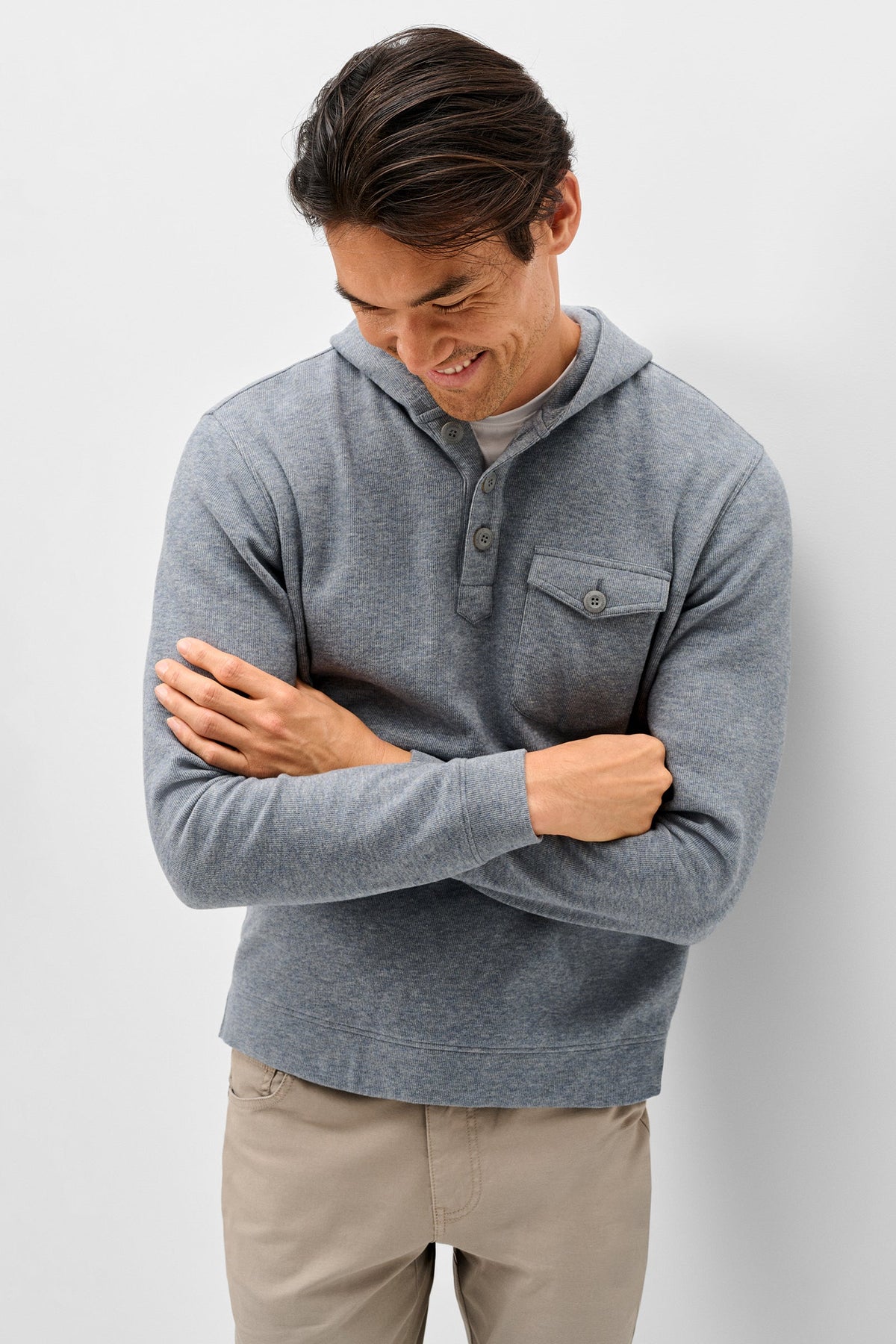 A man wearing a heather gray long-sleeve henley shirt with a hood and chest pocket, paired with beige pants, standing against a light gray studio background.