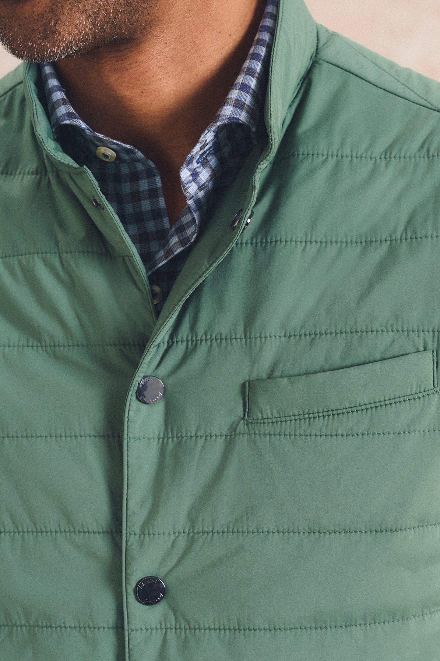 Close-up of a man wearing a sage green quilted puffer vest over a blue and white checkered button-down shirt.