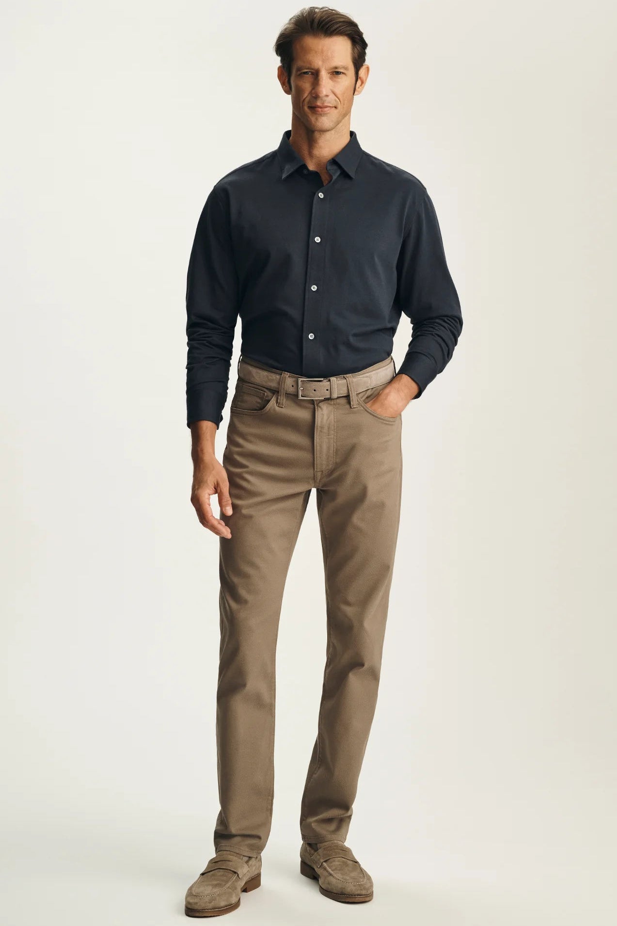 A man modeling a dark navy long-sleeve button-down shirt and tan chino pants with brown shoes, standing against a light beige studio background.