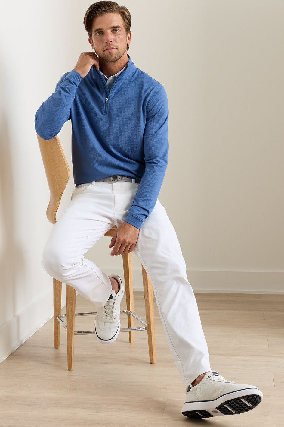 Man sitting on a stool wearing a blue sweater, white pants, and white sneakers.