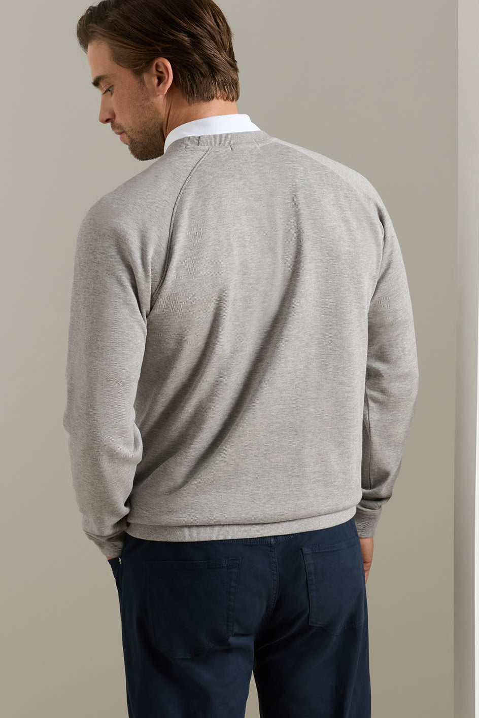 A man shown from the back wearing a light gray heather crewneck sweater over a white collared shirt and navy blue trousers, standing against a neutral beige wall.