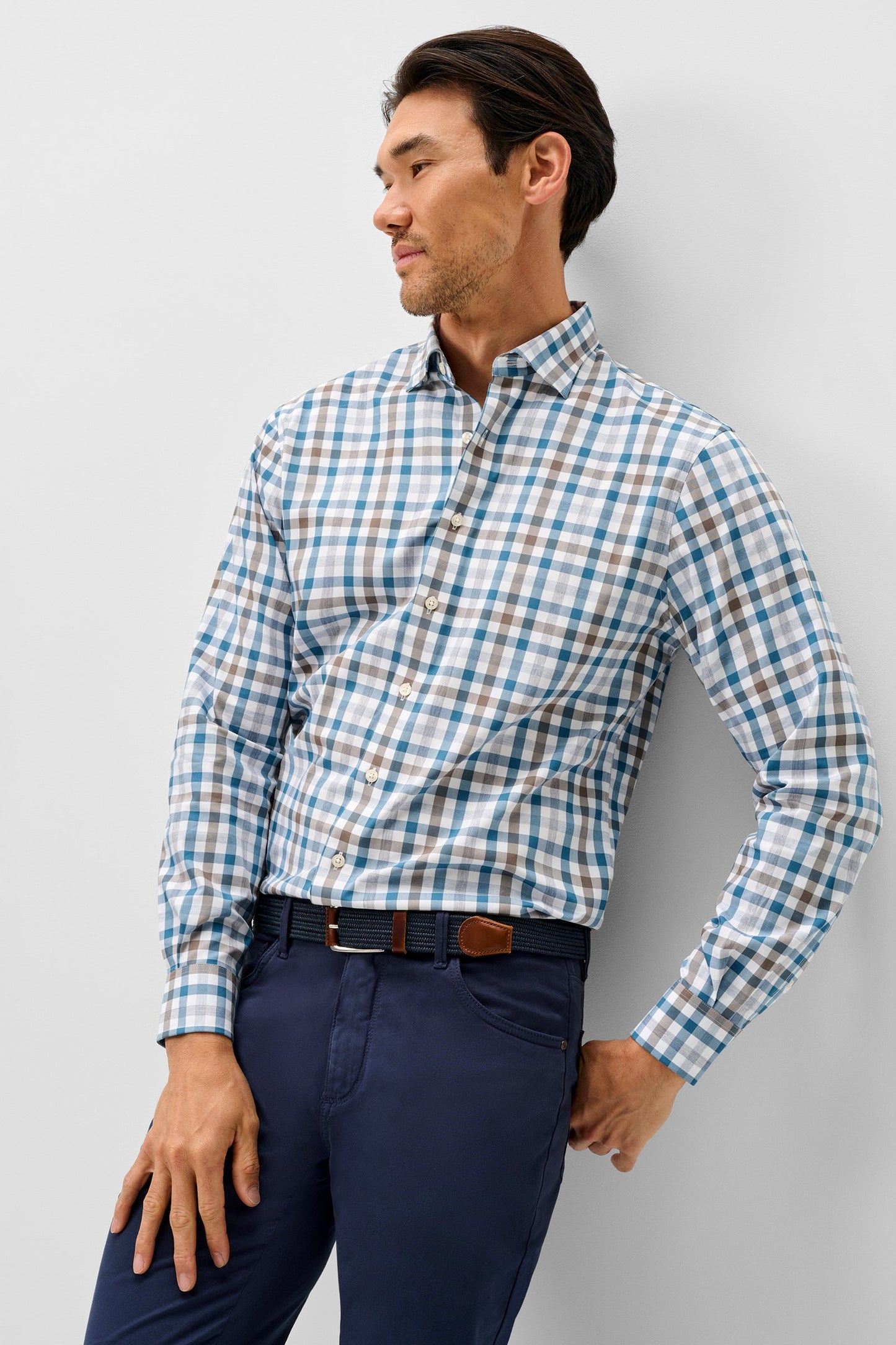 A man wearing a long-sleeve button-down shirt with a blue, white, and brown plaid pattern, paired with navy blue trousers and a brown belt, posing against a light gray wall.