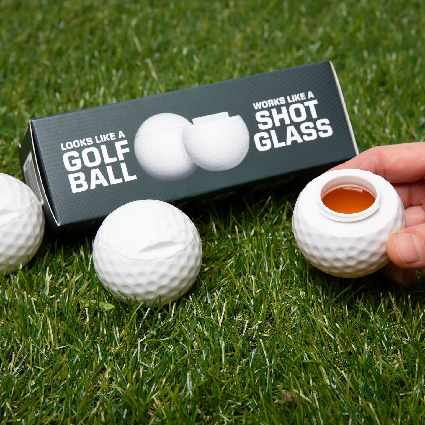 Golf ball-shaped shot glass on grass with packaging and additional golf balls.