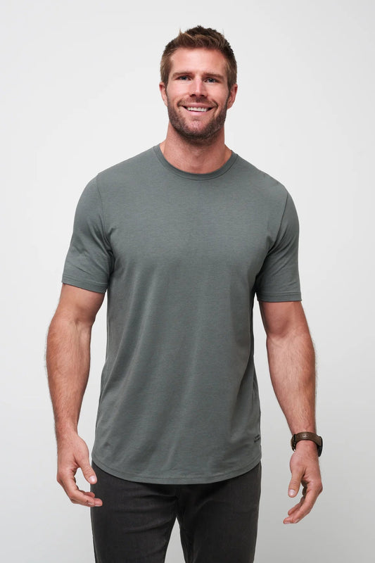 A smiling man wearing a short-sleeve crewneck t-shirt in a muted gray-green color, paired with dark pants, standing against a light gray studio background.