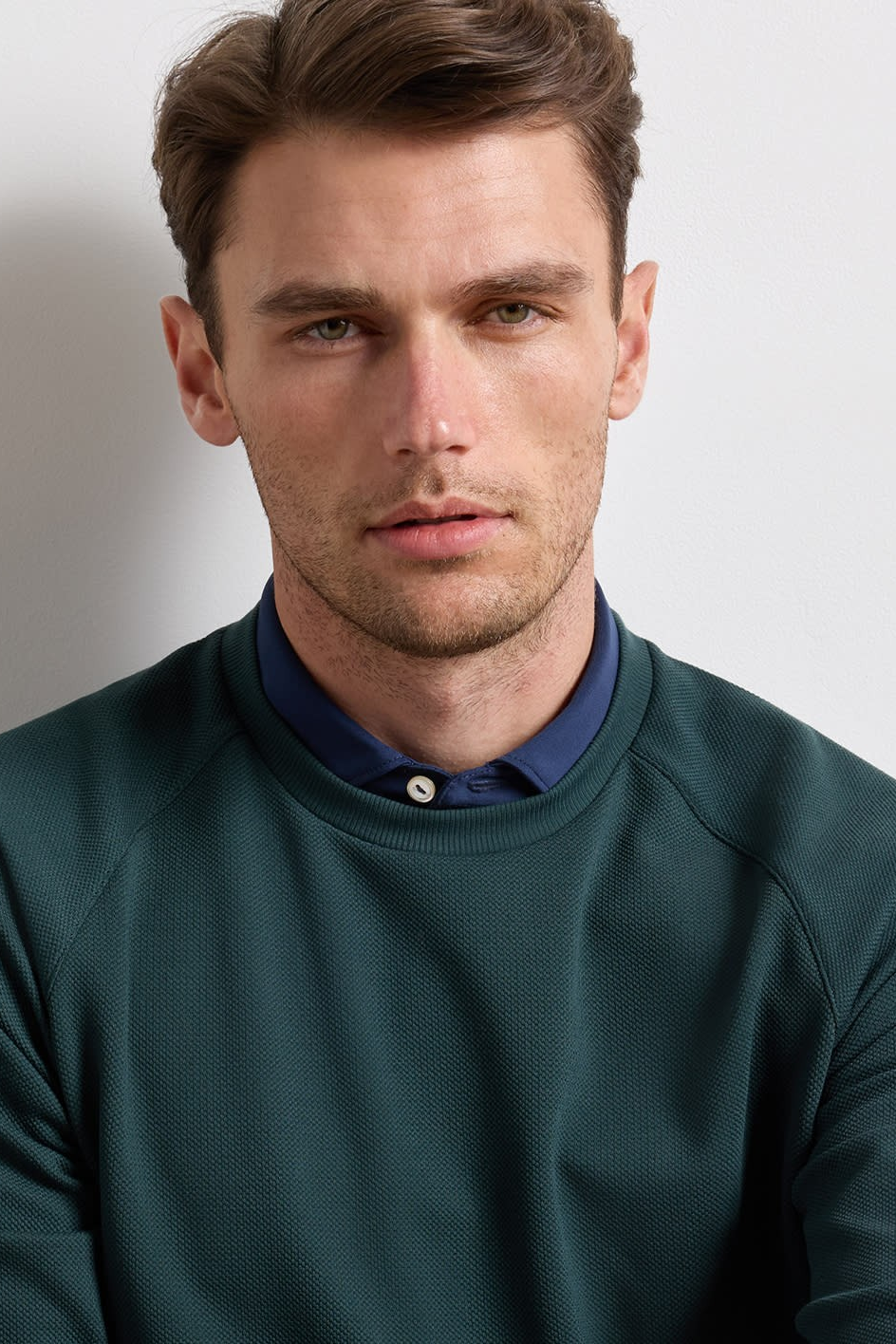 A man wearing a dark green crewneck sweatshirt layered over a navy blue button-down shirt, photographed against a plain light gray background.