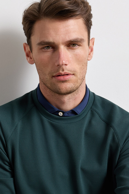 A man wearing a dark green crewneck sweatshirt layered over a navy blue button-down shirt, photographed against a plain light gray background.