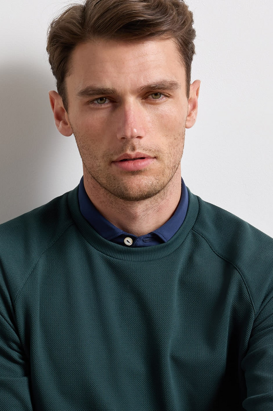 A man wearing a dark green crewneck sweatshirt layered over a navy blue button-down shirt, photographed against a plain light gray background.