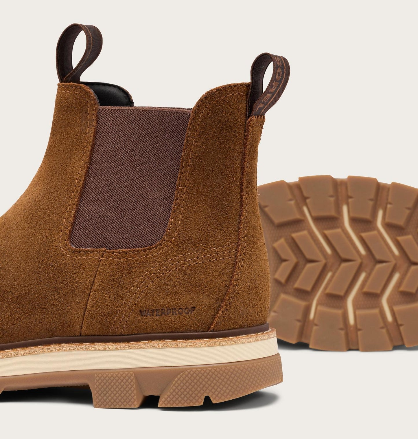 A close-up of a brown suede Chelsea boot with a textured brown sole and a separate outsole shown behind it, on a light beige background.