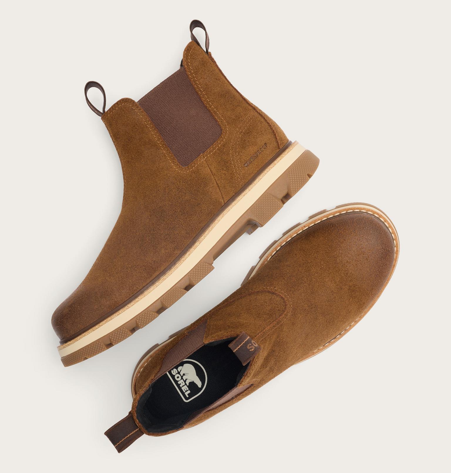 A pair of brown suede Chelsea-style boots shown from top and side views on a light gray background. One boot shows a dark brown elastic side panel, a tan midsole, and pull tabs.