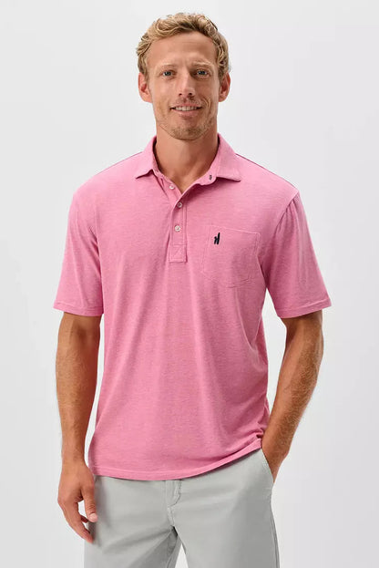 A man wearing a pink short-sleeve polo shirt with a small dark embroidered logo on the left chest, paired with light gray trousers, photographed against a clean light gray studio background.