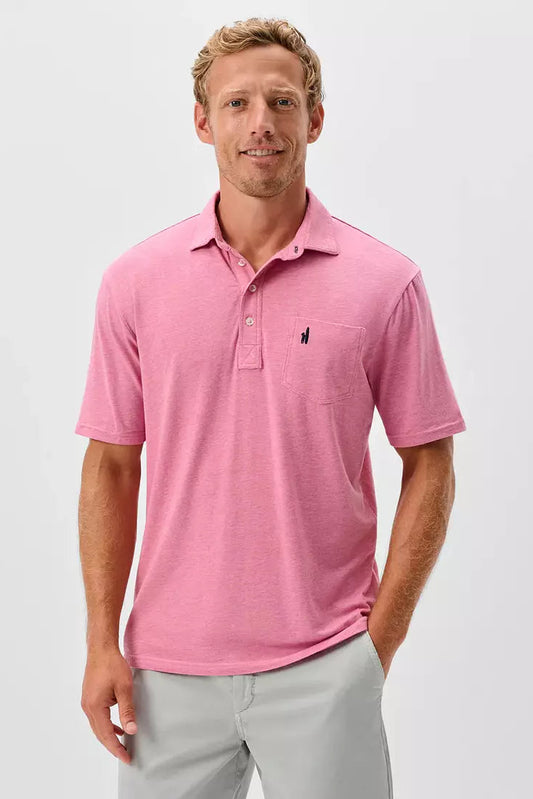 A man wearing a pink short-sleeve polo shirt with a small dark embroidered logo on the left chest, paired with light gray trousers, photographed against a clean light gray studio background.