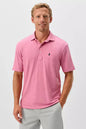 A man wearing a pink short-sleeve polo shirt with a small dark embroidered logo on the left chest, paired with light gray trousers, photographed against a clean light gray studio background.
