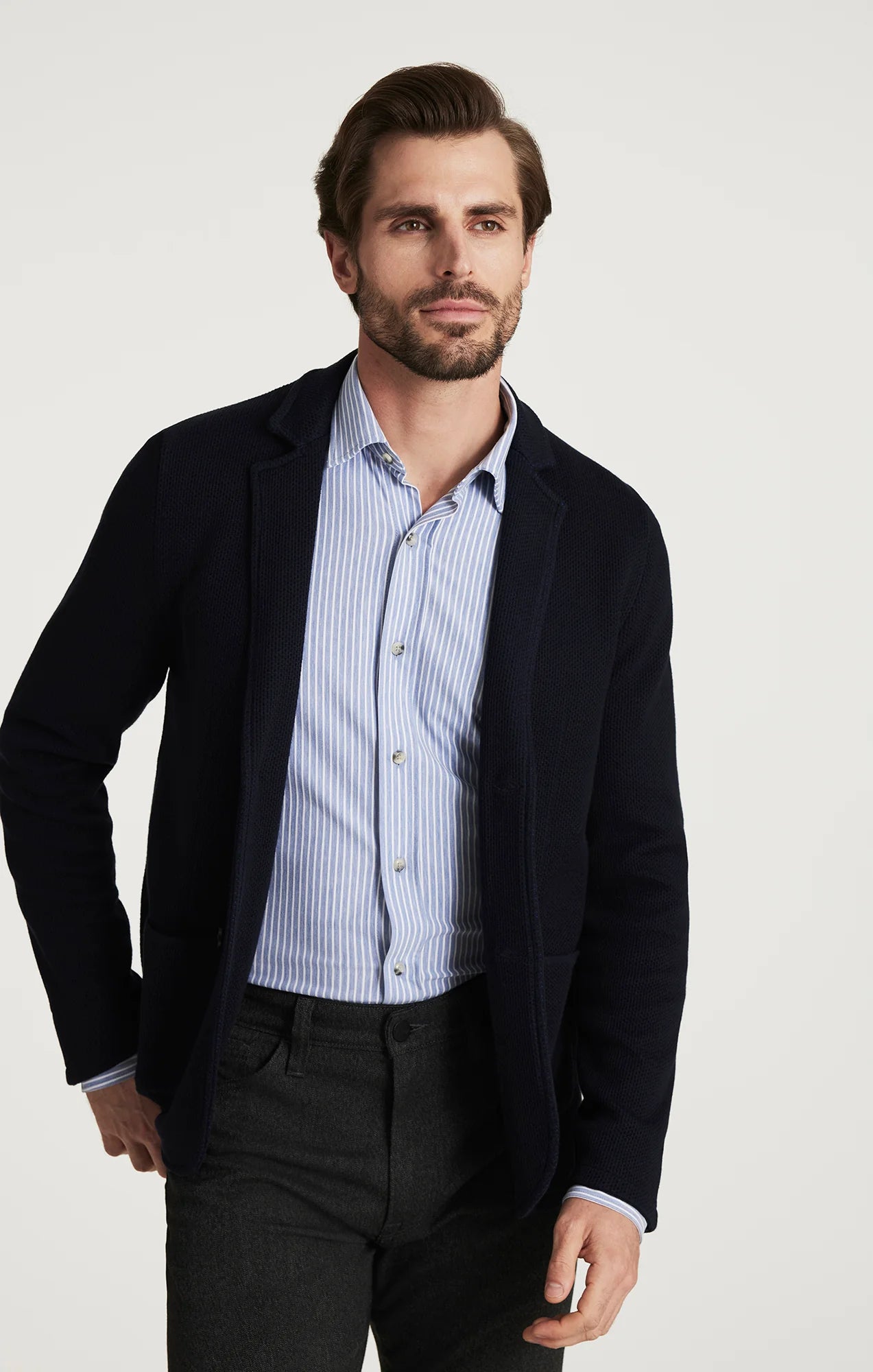 A man wearing a dark navy blazer over a light blue and white striped button-down shirt, paired with dark charcoal trousers, photographed against a clean off-white studio background.