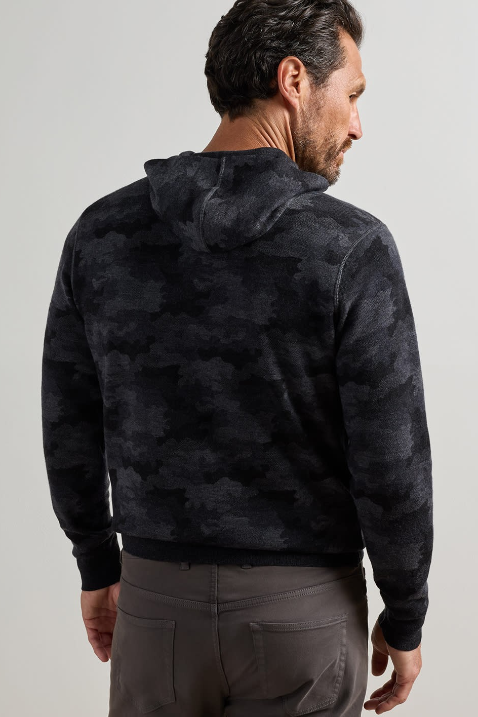A man wearing a dark charcoal gray hooded sweatshirt with a subtle tonal camouflage pattern, shown from the back against a light gray studio background.
