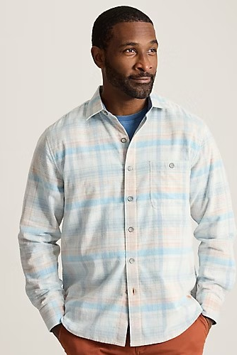 A man wearing a long-sleeve button-up plaid shirt in light blue, white, and beige, layered over a blue t-shirt and paired with rust-orange pants, against a solid light gray background.