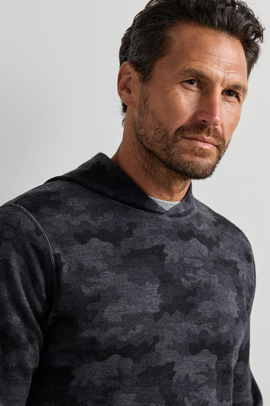 A close-up of a man wearing a dark gray and black camouflage hoodie sweatshirt, photographed against a neutral light gray background.