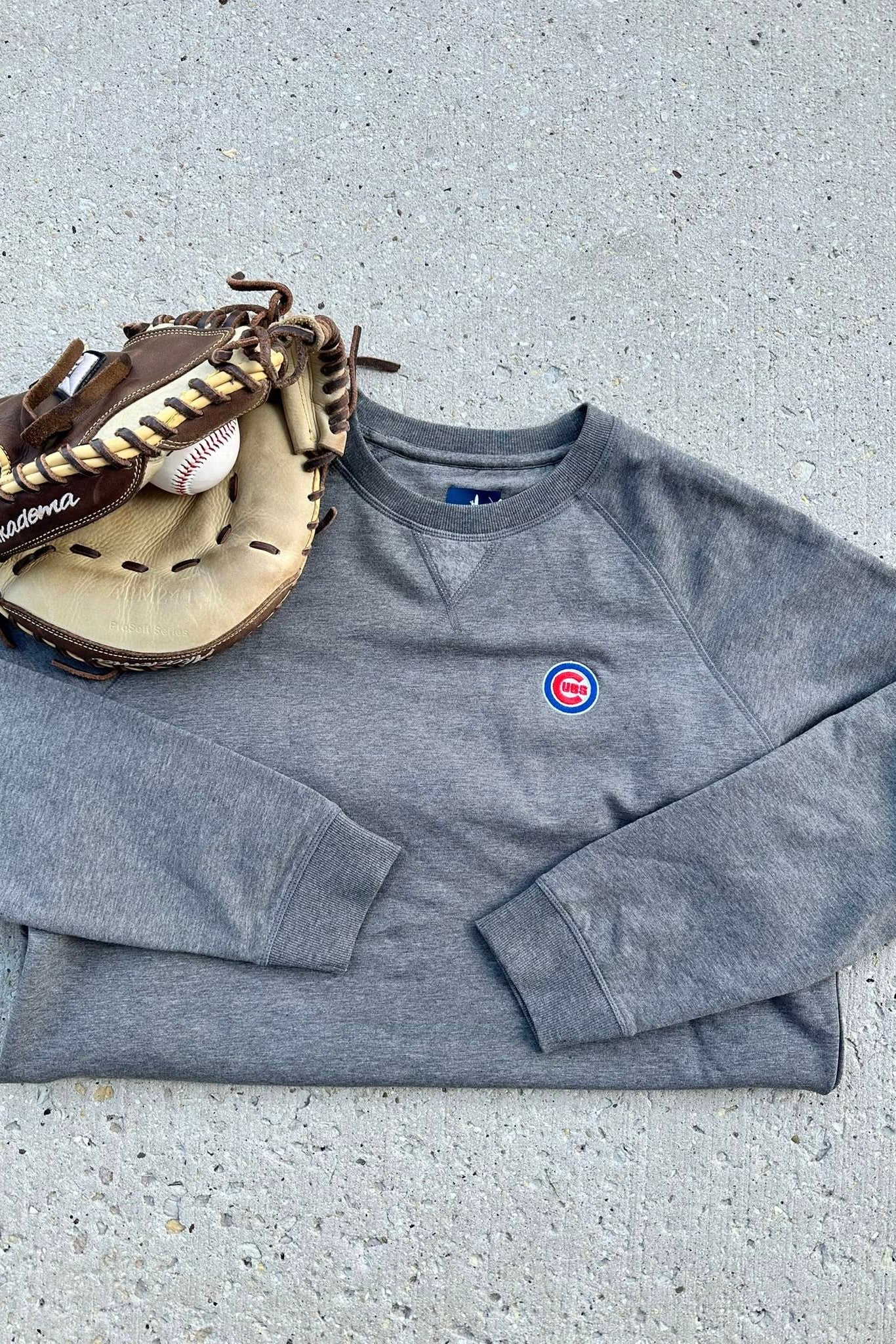 Gray sweatshirt with a logo, baseball glove, and ball on a concrete surface