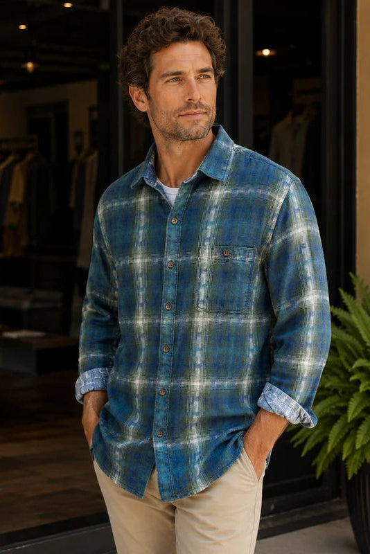A man wearing a blue and green plaid button-down flannel shirt with beige pants, standing in front of a storefront with a green fern plant in the background.