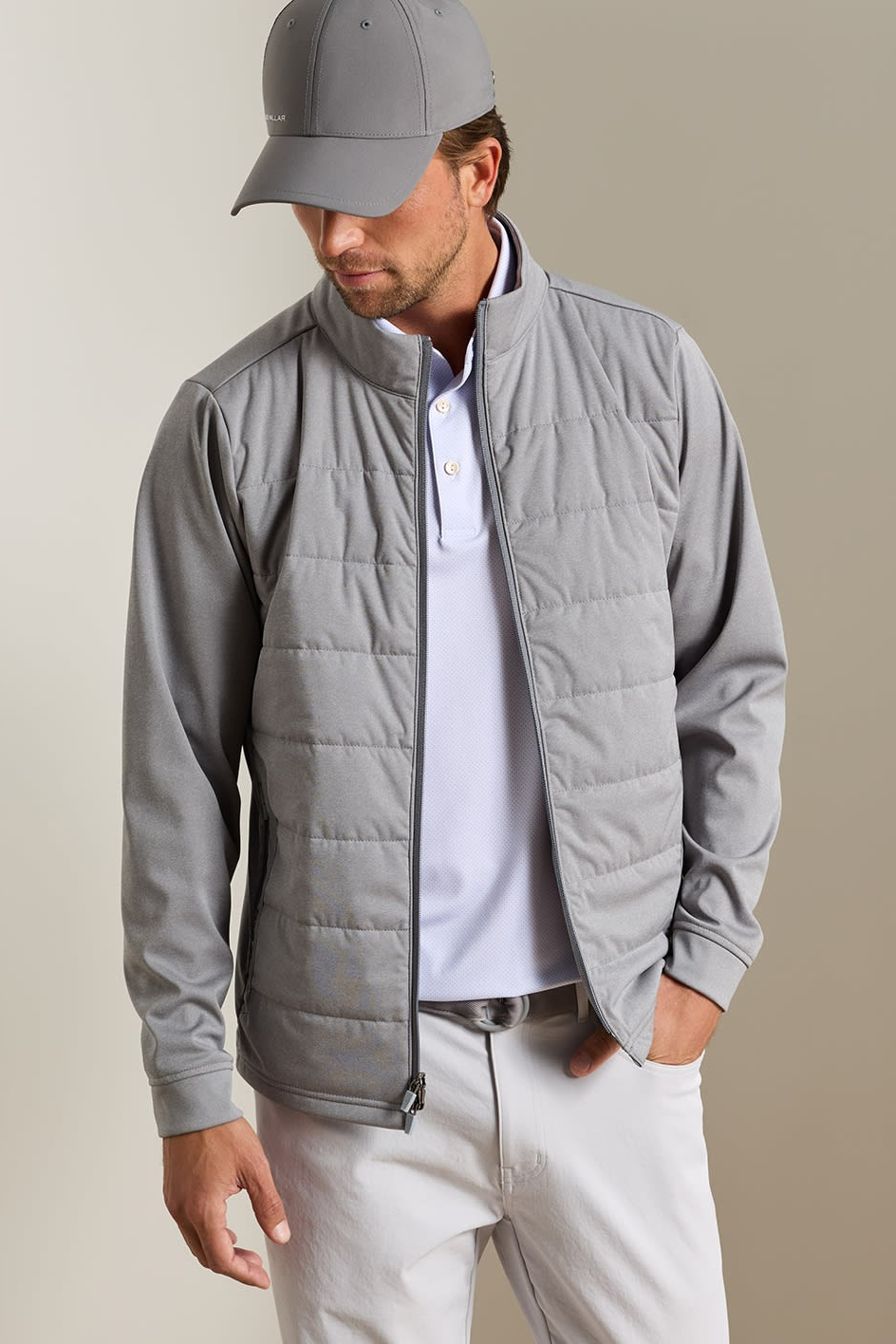 A man wearing a light gray zip-up puffer jacket over a white polo shirt, paired with a gray baseball cap and off-white trousers, standing against a neutral beige background.