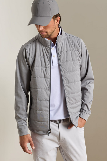 A man wearing a light gray zip-up puffer jacket over a white polo shirt, paired with a gray baseball cap and off-white trousers, standing against a neutral beige background.