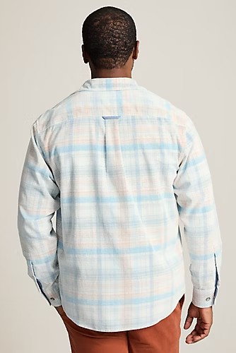 A man viewed from behind wearing a long-sleeve plaid button-up shirt in white, light blue, and pale pink, paired with rust-brown pants, against a solid light beige background.