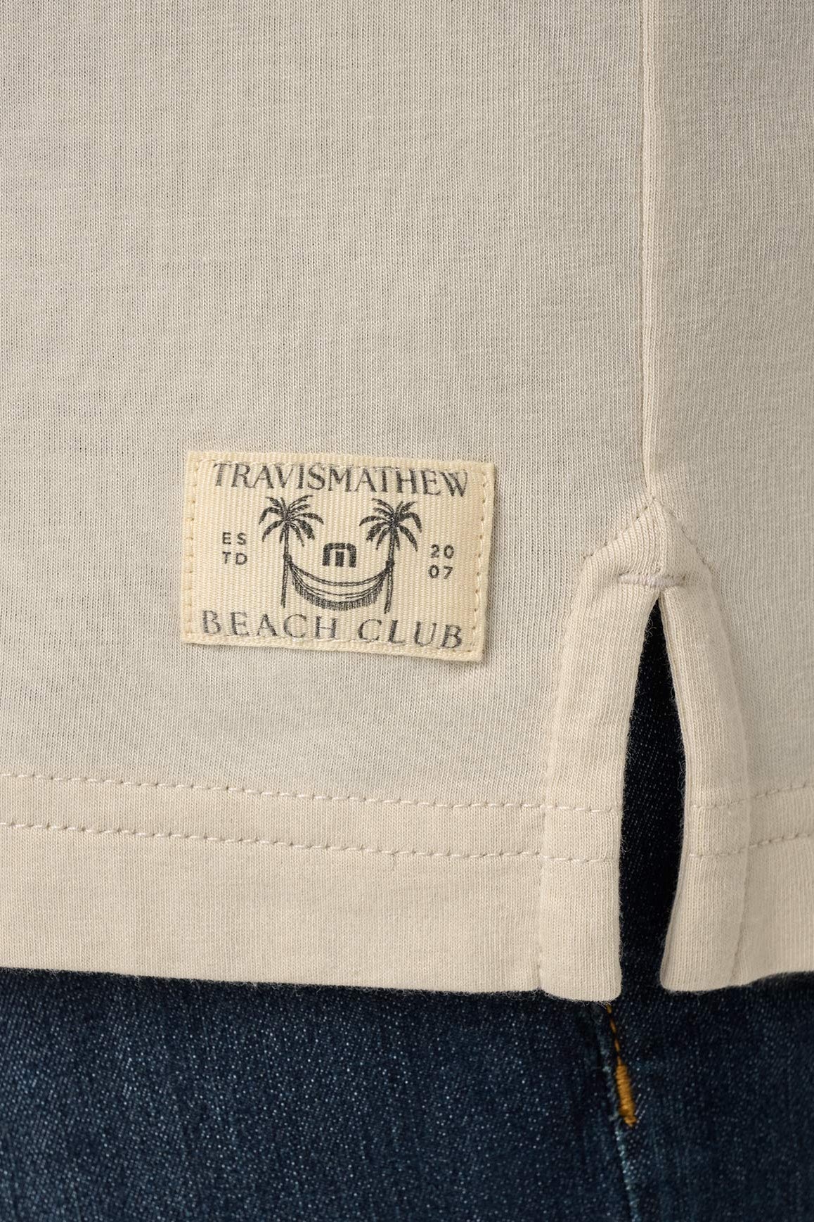 Close-up of a label on a garment with 'Travismathew Beach Club' text and palm tree design.