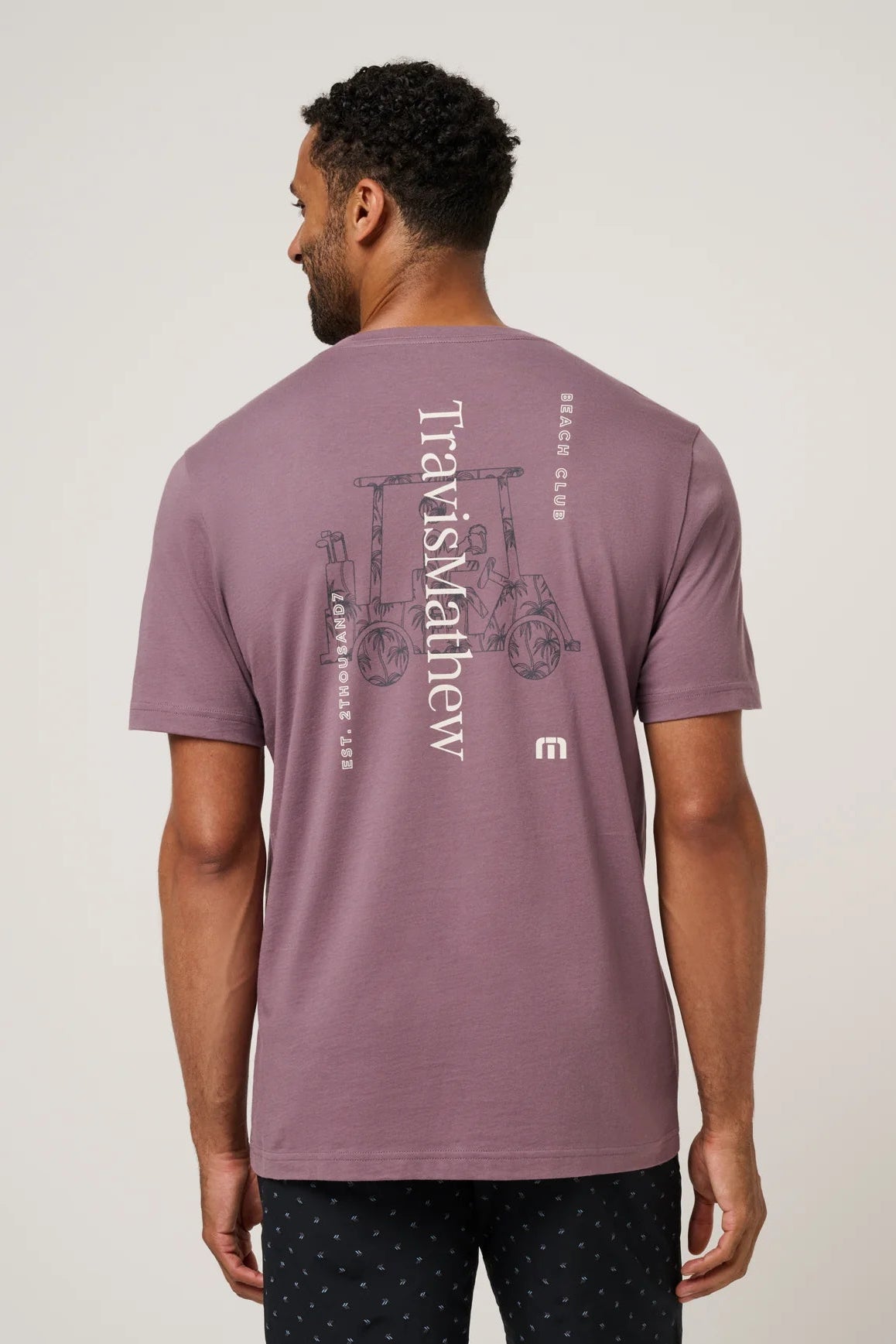 Man wearing a purple t-shirt with 'Travis Mathew' branding on the back, standing against a plain background.