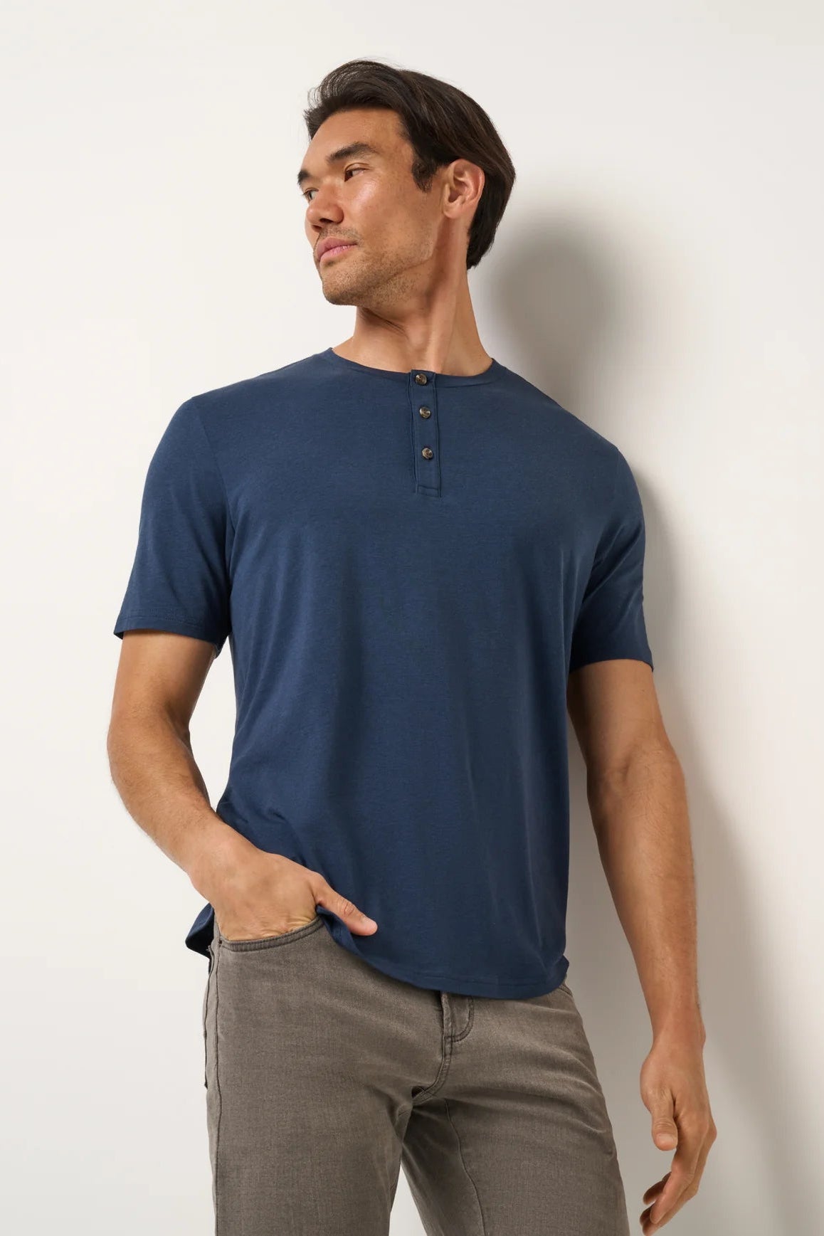 Man wearing a blue henley shirt and gray pants against a white wall