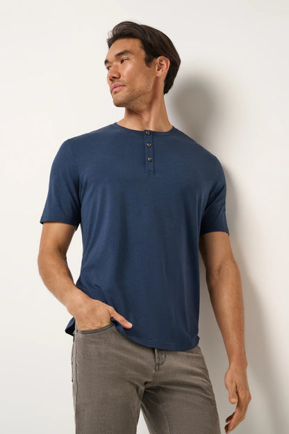 Man wearing a blue henley shirt and gray pants against a white wall