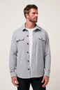 A man wearing a light gray long-sleeve button-up overshirt over a white t-shirt and dark jeans, standing against a plain light gray studio background.