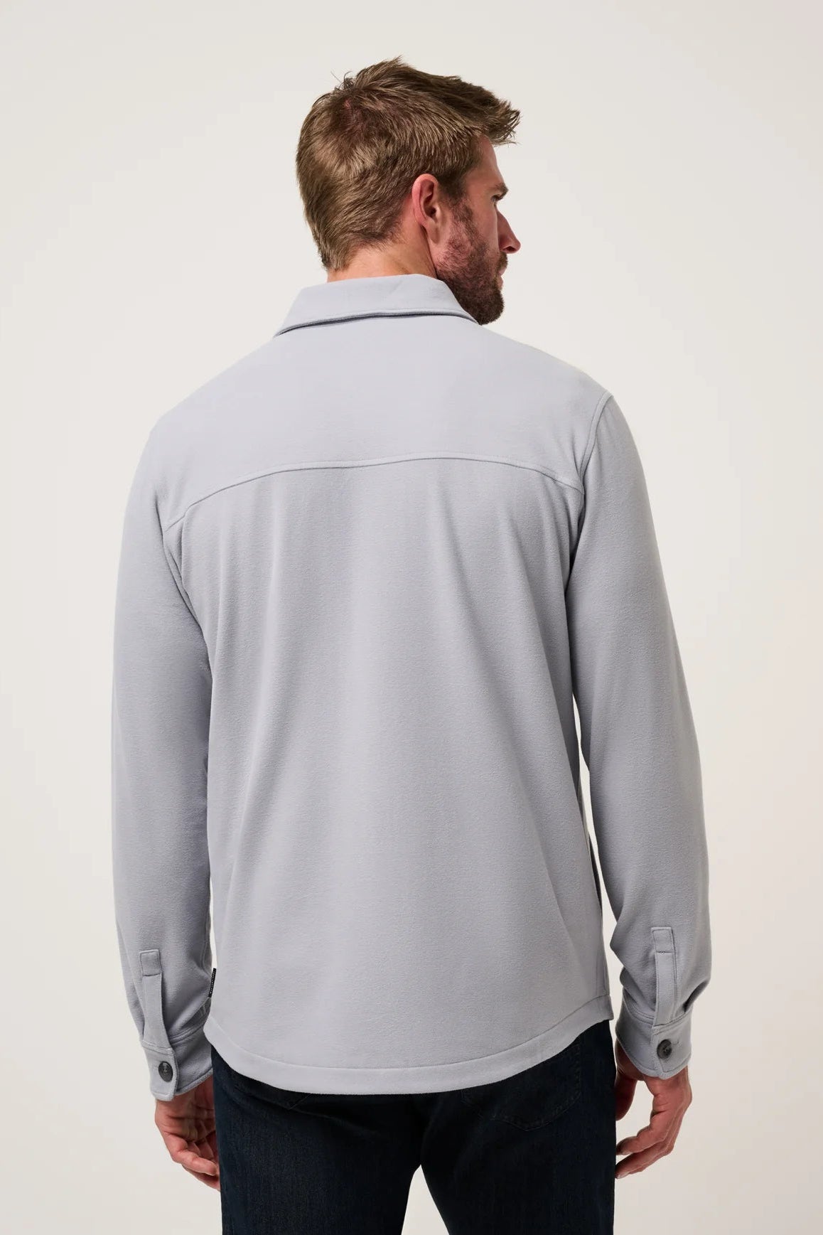 A man viewed from the back wearing a light gray long-sleeve button-up shirt and dark jeans, standing against a plain off-white studio background.