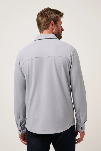 A man viewed from the back wearing a light gray long-sleeve button-up shirt and dark jeans, standing against a plain off-white studio background.
