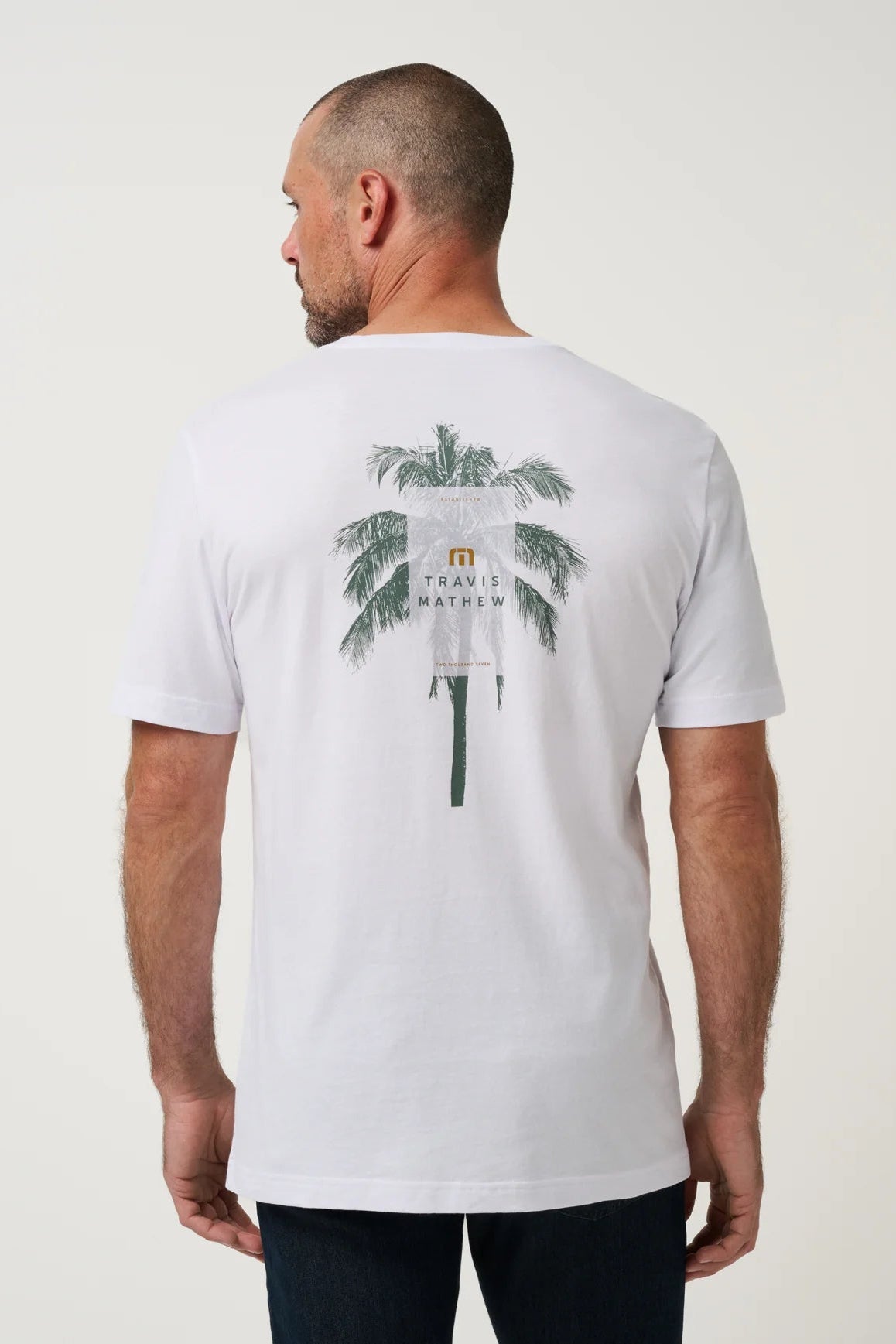 A man viewed from the back wearing a white short-sleeve t-shirt with a large graphic print of a palm tree on the back. The graphic features a white flag with the text 'TRAVEL MEXICO' and a small yellow emblem.