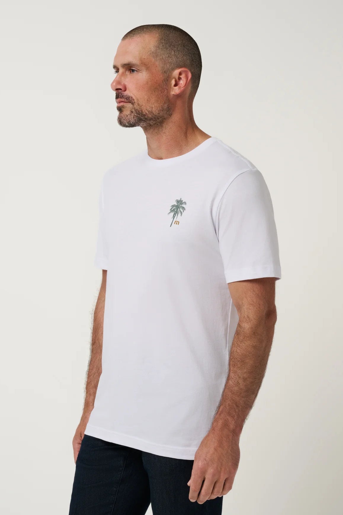 A man wearing a white short-sleeve crewneck t-shirt with a small green palm tree graphic on the left chest, paired with dark pants, photographed against a plain light background.