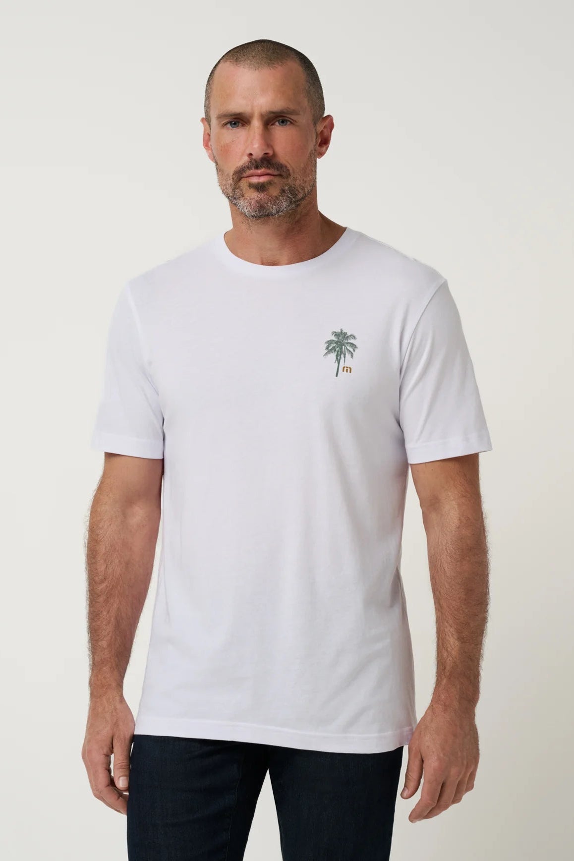 A man wearing a white short-sleeve crewneck t-shirt with a small green palm tree graphic on the left chest, paired with dark pants, against a plain light gray studio background.