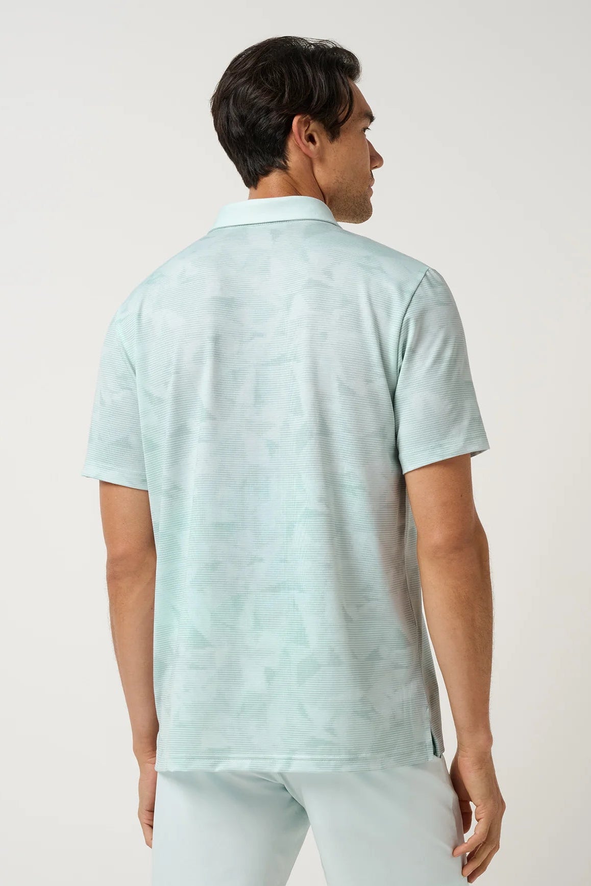 A man viewed from the back wearing a short-sleeve polo shirt in a very light mint green color with a subtle tonal pattern, paired with matching light pants, against a plain off-white studio background.