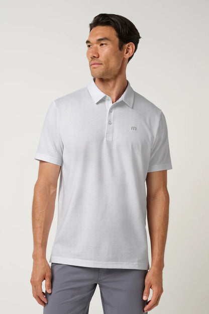 A man wearing a light gray short-sleeve polo shirt with a small embroidered logo on the left chest, paired with gray trousers, photographed against a plain light gray studio background.