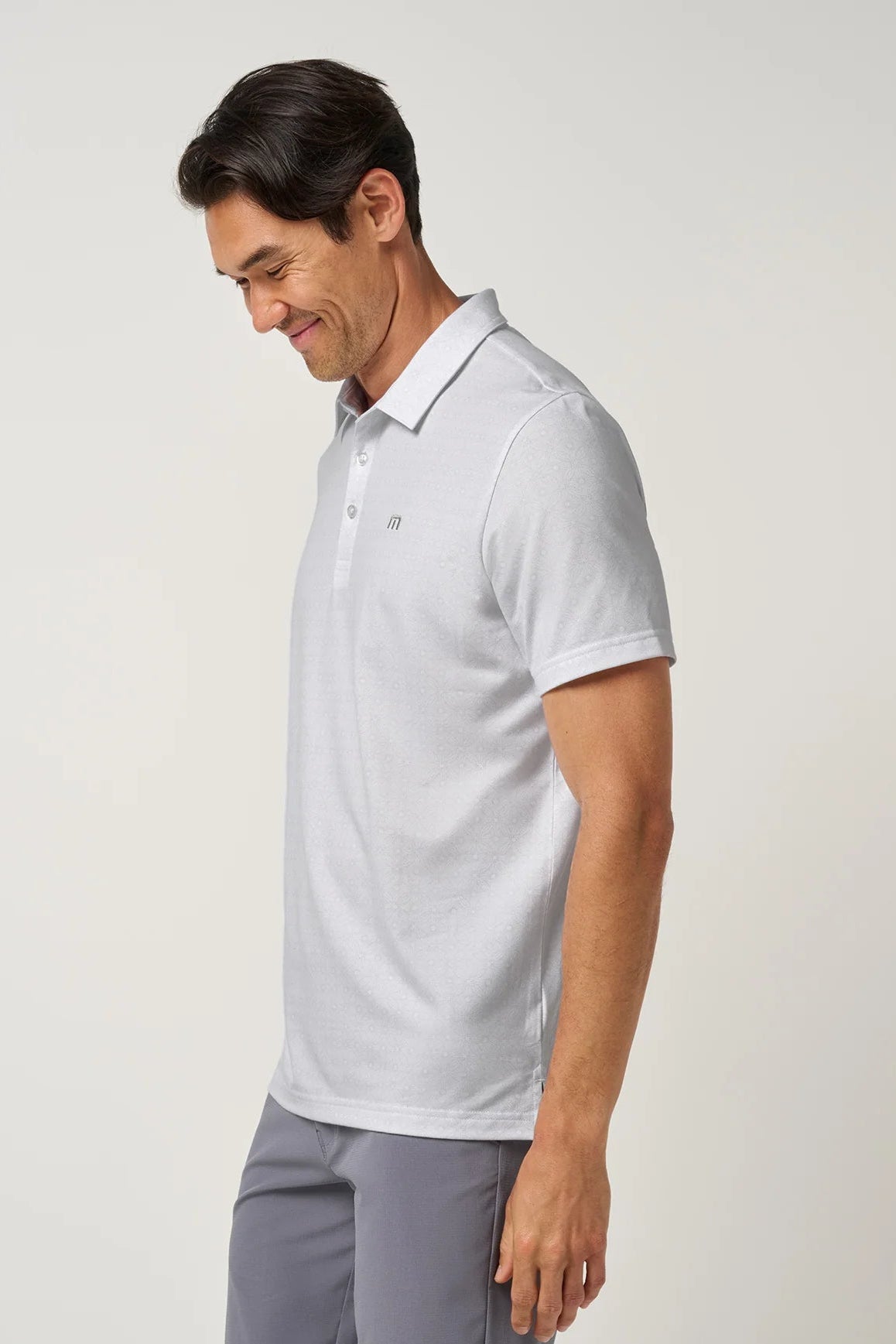 A man wearing a light gray short-sleeve polo shirt and gray trousers, shown in profile against a plain light gray studio background.