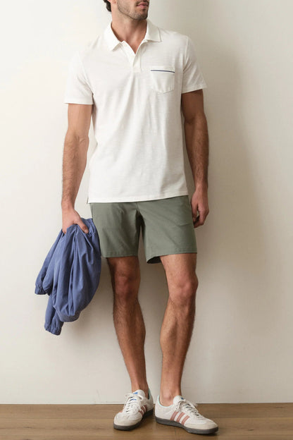 Man wearing a white polo shirt, green shorts, and white sneakers, holding a blue jacket against a plain background.