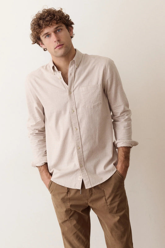 A man wearing a light beige linen long-sleeve button-down shirt and brown trousers, standing against a plain off-white background.