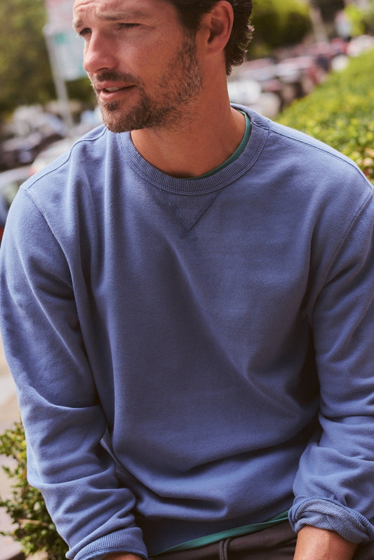 A man wearing a dusty blue crewneck sweatshirt, sitting outdoors on a stone ledge with green bushes and a blurred street background.