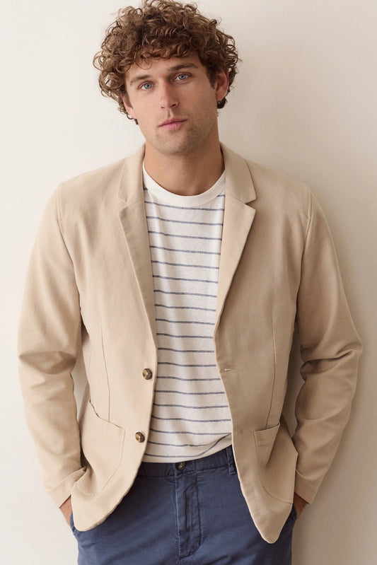 A man wearing a beige blazer over a white striped t-shirt and blue jeans, standing against a plain beige wall.