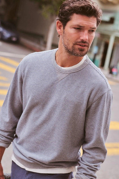 A man wearing a light gray ribbed crewneck sweatshirt over a white undershirt, standing on an outdoor street with blurred buildings in the background.