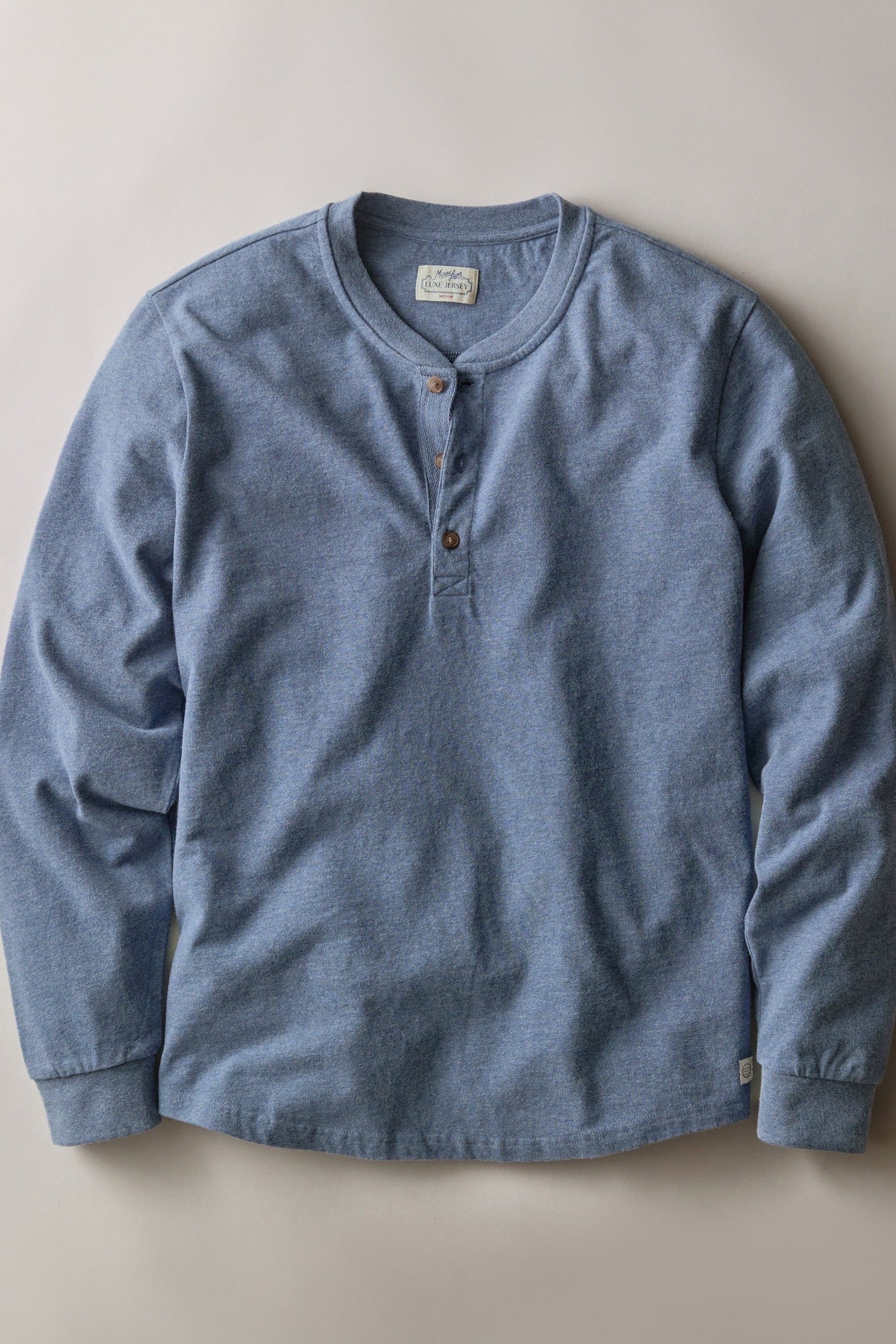A light blue heathered long-sleeve henley shirt with a three-button placket, laid flat against a neutral beige background.