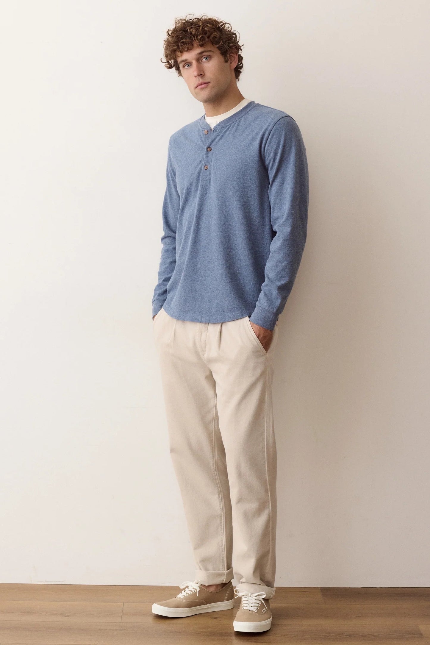 A man wearing a dusty blue long-sleeve henley shirt, beige trousers, and beige sneakers, standing against a plain off-white wall.