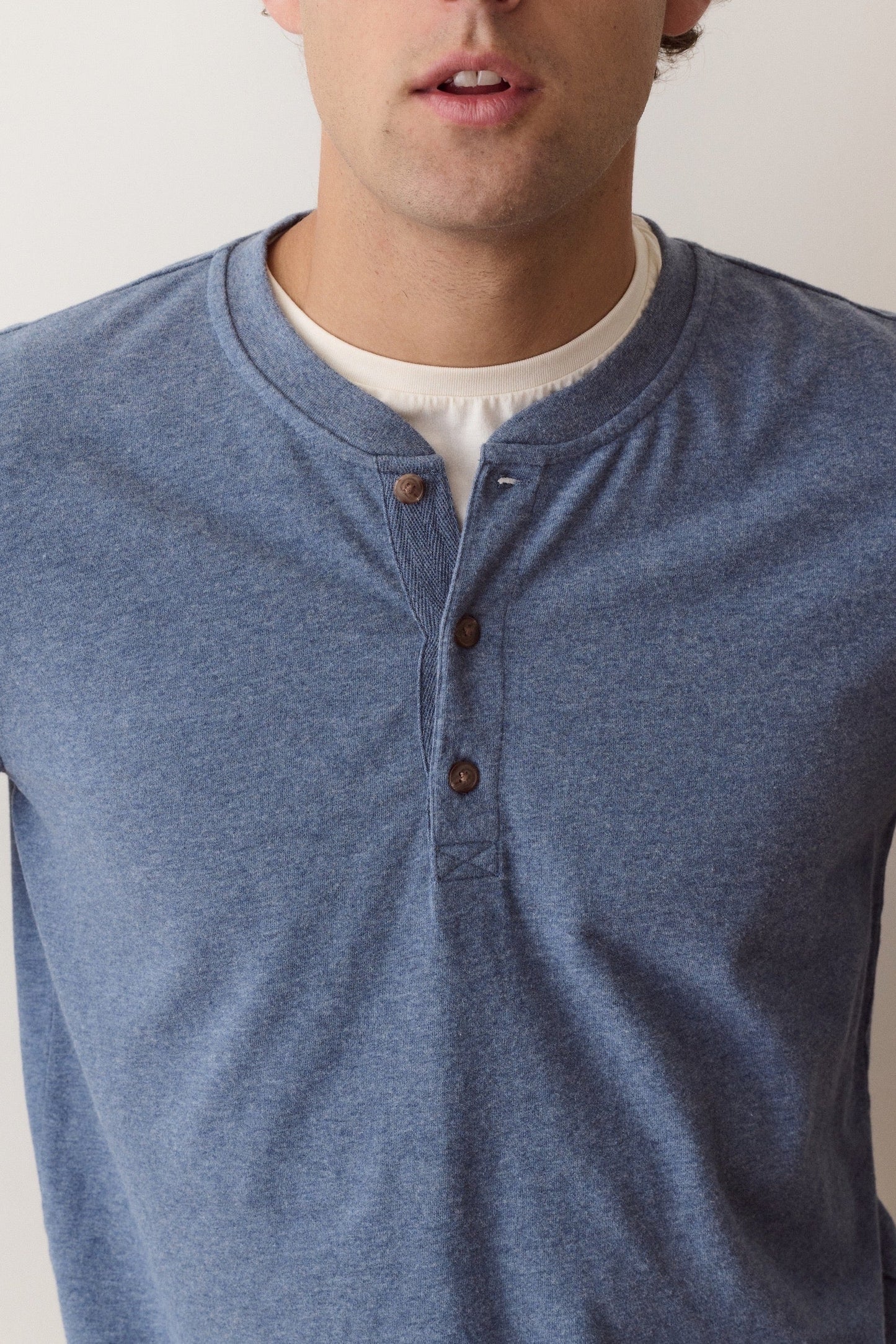 A man wearing a heathered blue long-sleeve henley shirt over a white undershirt, shown from the chest up against a plain light background.