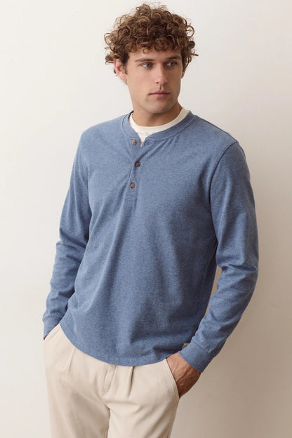 A man wearing a heathered blue long-sleeve henley shirt with brown buttons over a white undershirt and beige trousers, standing against a plain off-white background.