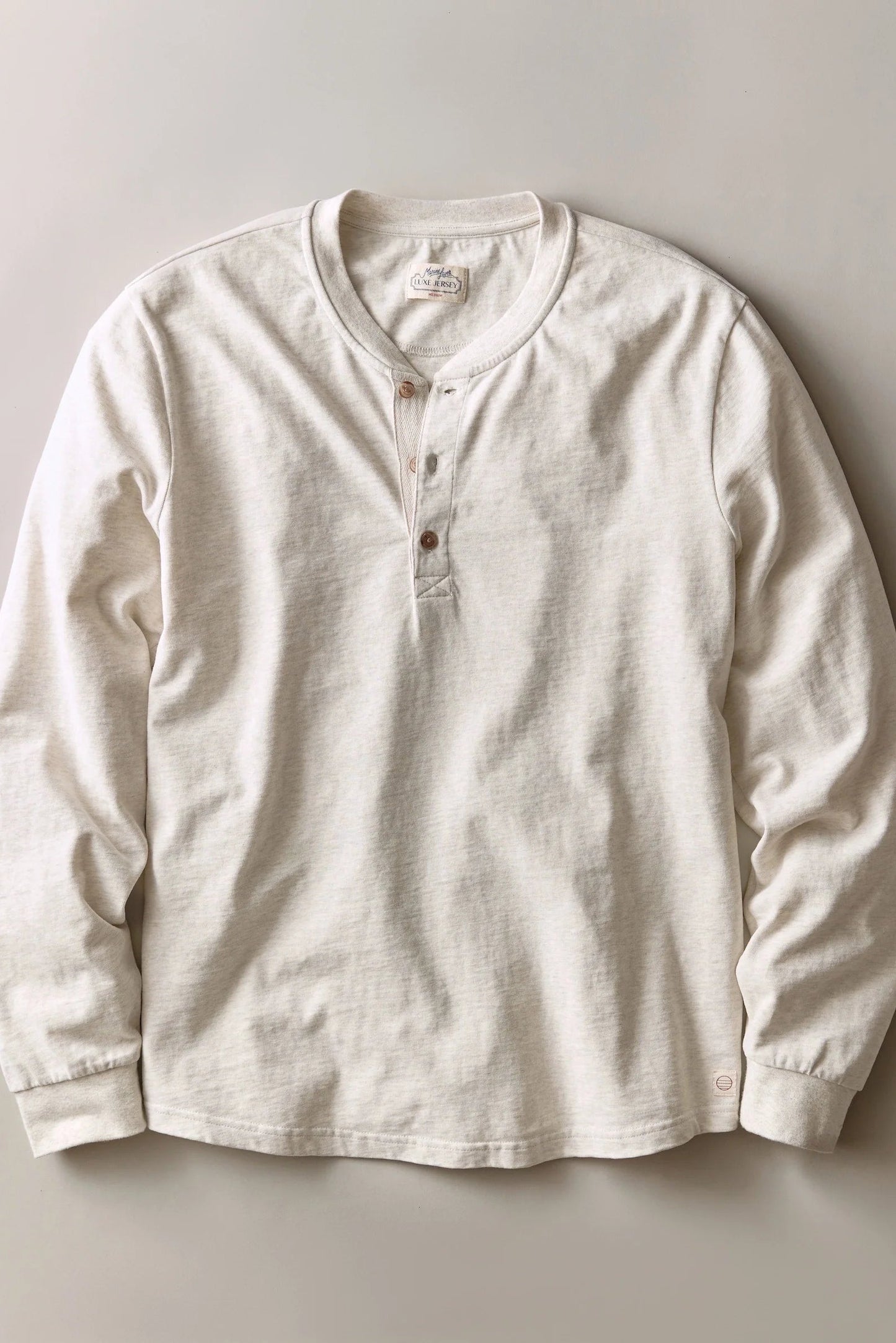 An off-white long-sleeve henley shirt with a three-button placket, laid flat against a neutral light gray background.