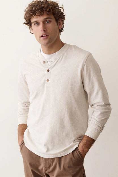 A man wearing a light cream long-sleeve henley shirt and brown pants, standing against a plain off-white background.