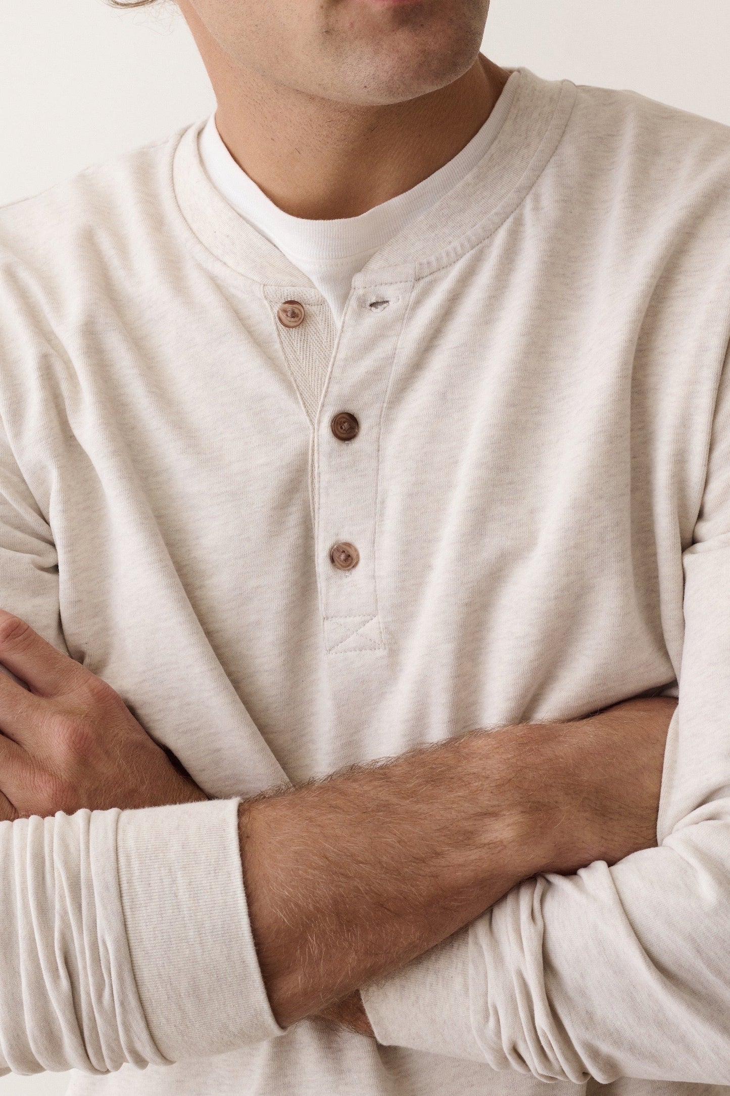 A close-up of a man wearing a light beige long-sleeve henley shirt with brown buttons, layered over a white undershirt, with his arms crossed.